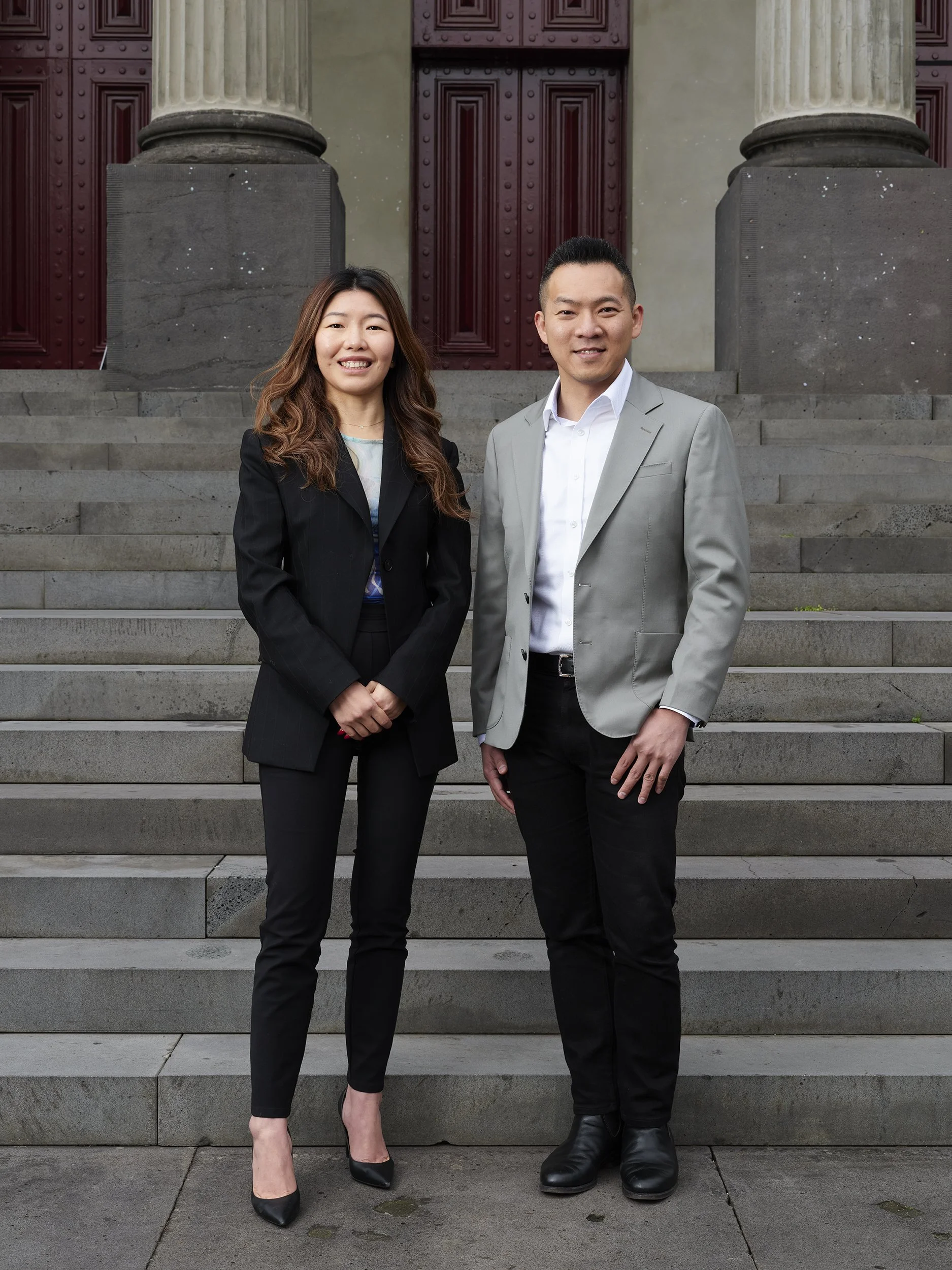 Two professionally dressed individuals, a woman and a man, standing on the steps in front of a building with large columns and red double doors.