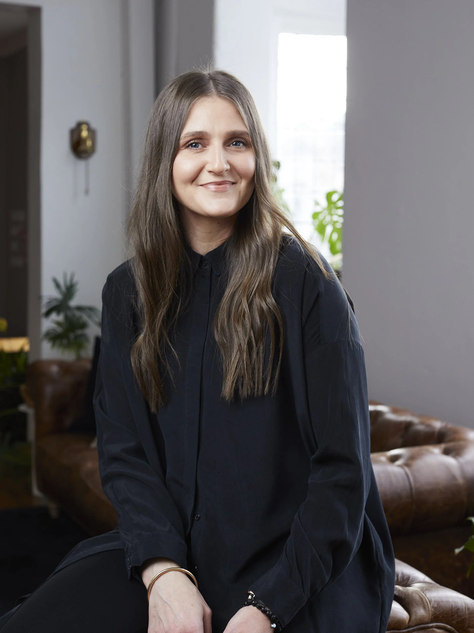 A young woman with long brown hair wearing a black shirt, smiling while sitting on a brown leather couch in a modern, well-lit room with plants and large windows.