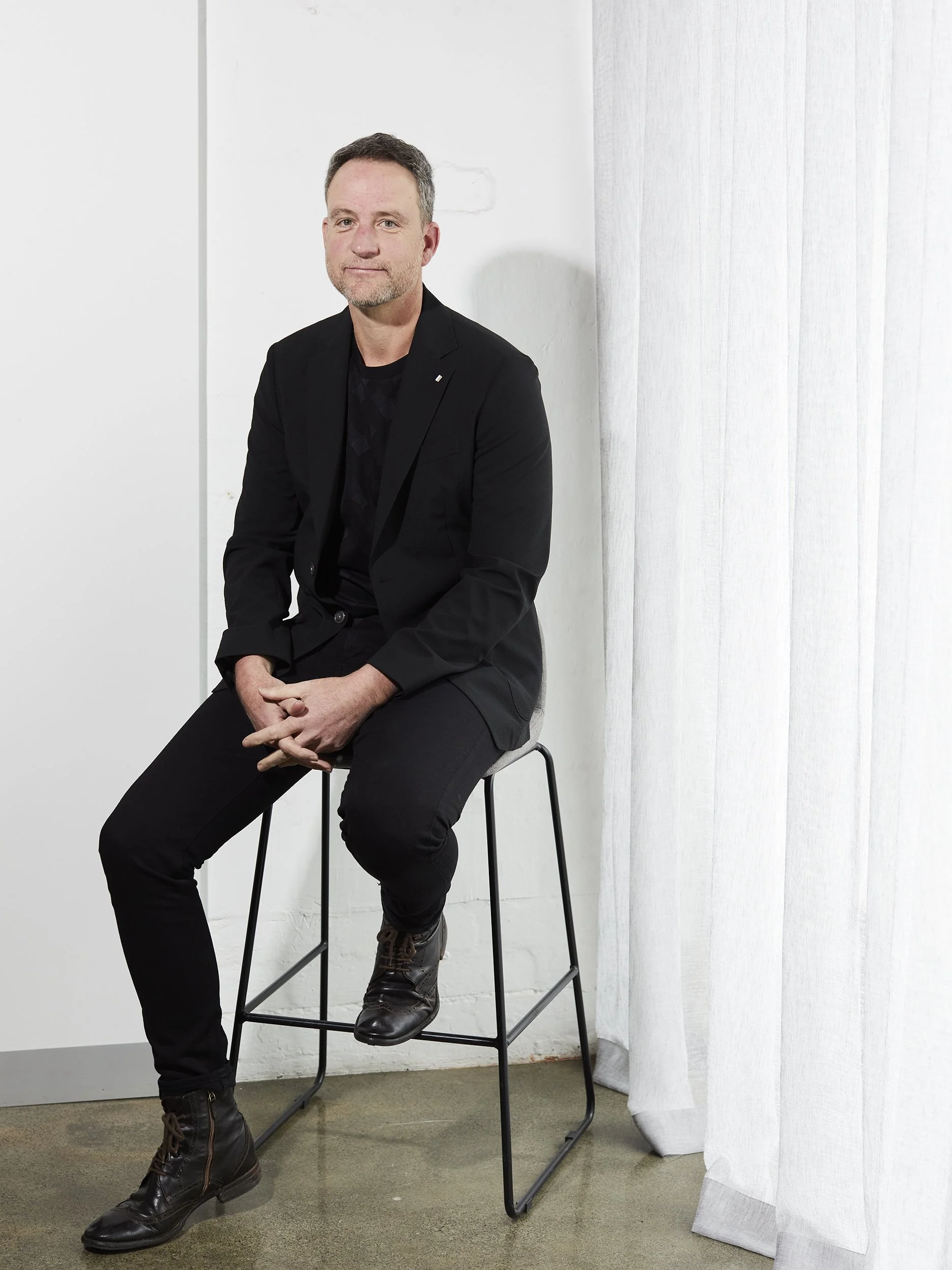 A man with short dark hair and a beard, dressed in black, sitting on a high stool in a minimalistic white room with a white curtain.