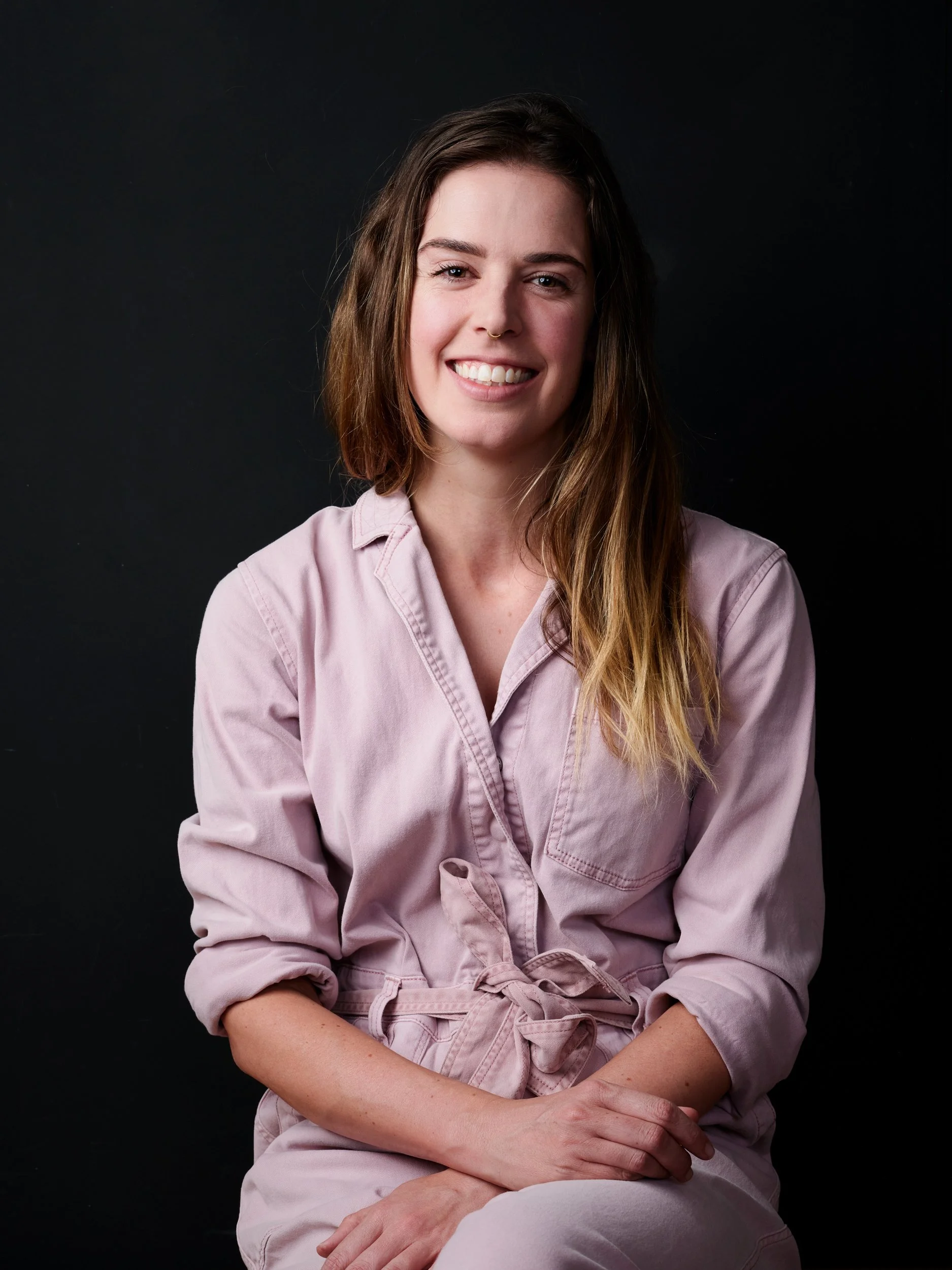 A woman with long brown hair, smiling, wearing a light pink jacket with rolled-up sleeves, seated against a dark background.