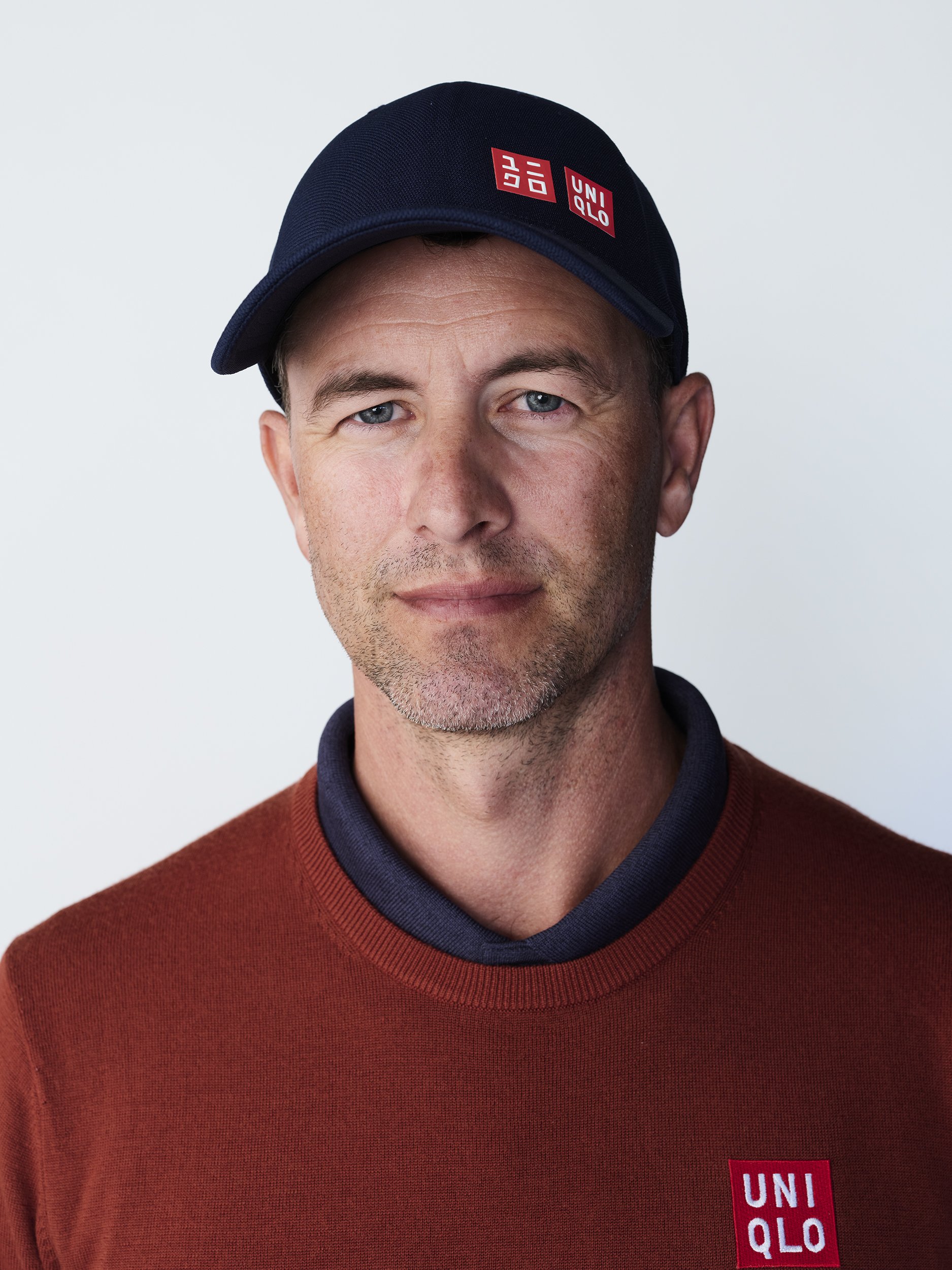 A man with light skin, blue eyes, and short brown hair wearing a navy blue cap with the Uniqlo logo, a dark blue collar shirt, and a red sweater with the Uniqlo logo.