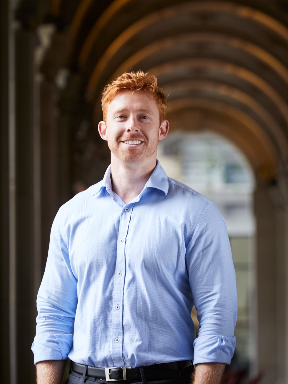 A man with red hair, wearing a light blue dress shirt, standing under a series of arched structures, smiling at the camera.