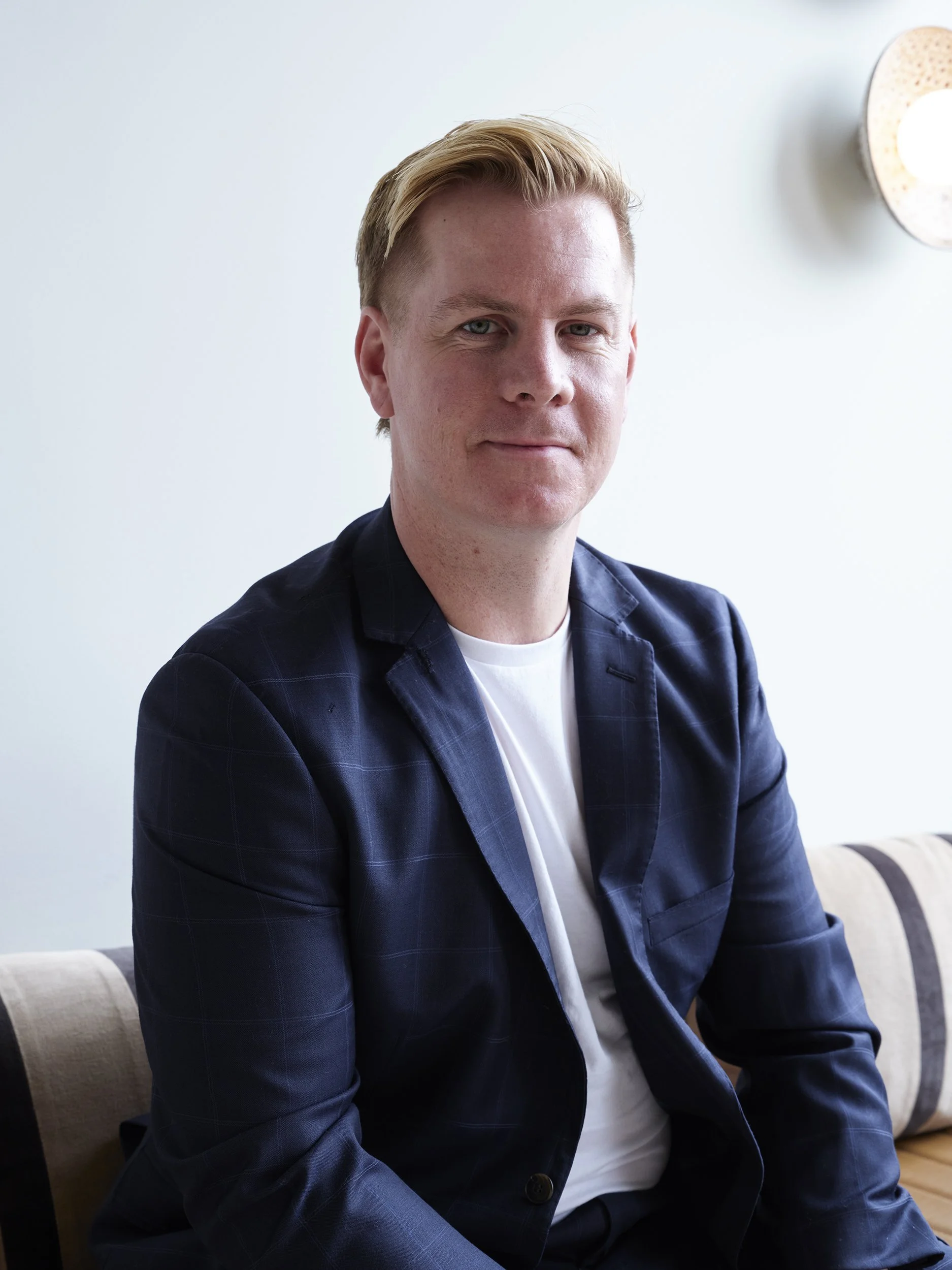 A young man with blonde hair and fair skin, wearing a navy blazer and white t-shirt, sitting on a beige and black striped sofa in a modern indoor setting.