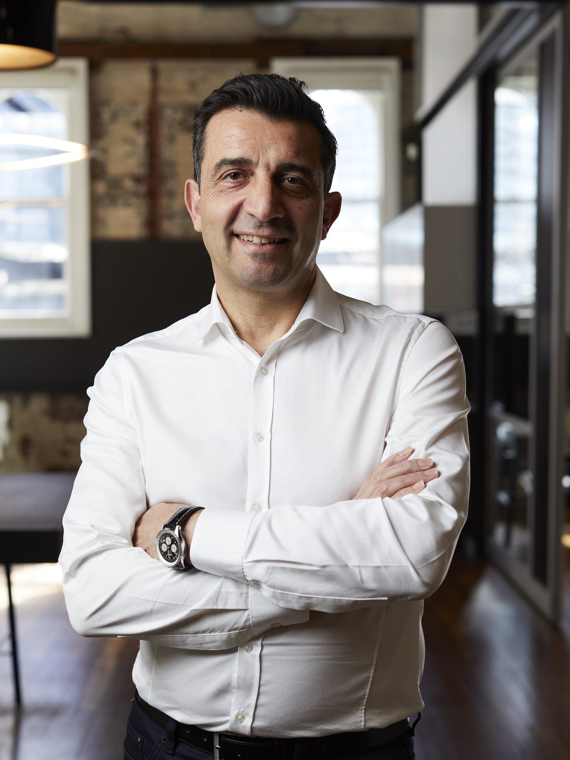A smiling man in a white shirt standing with arms crossed in a modern office.