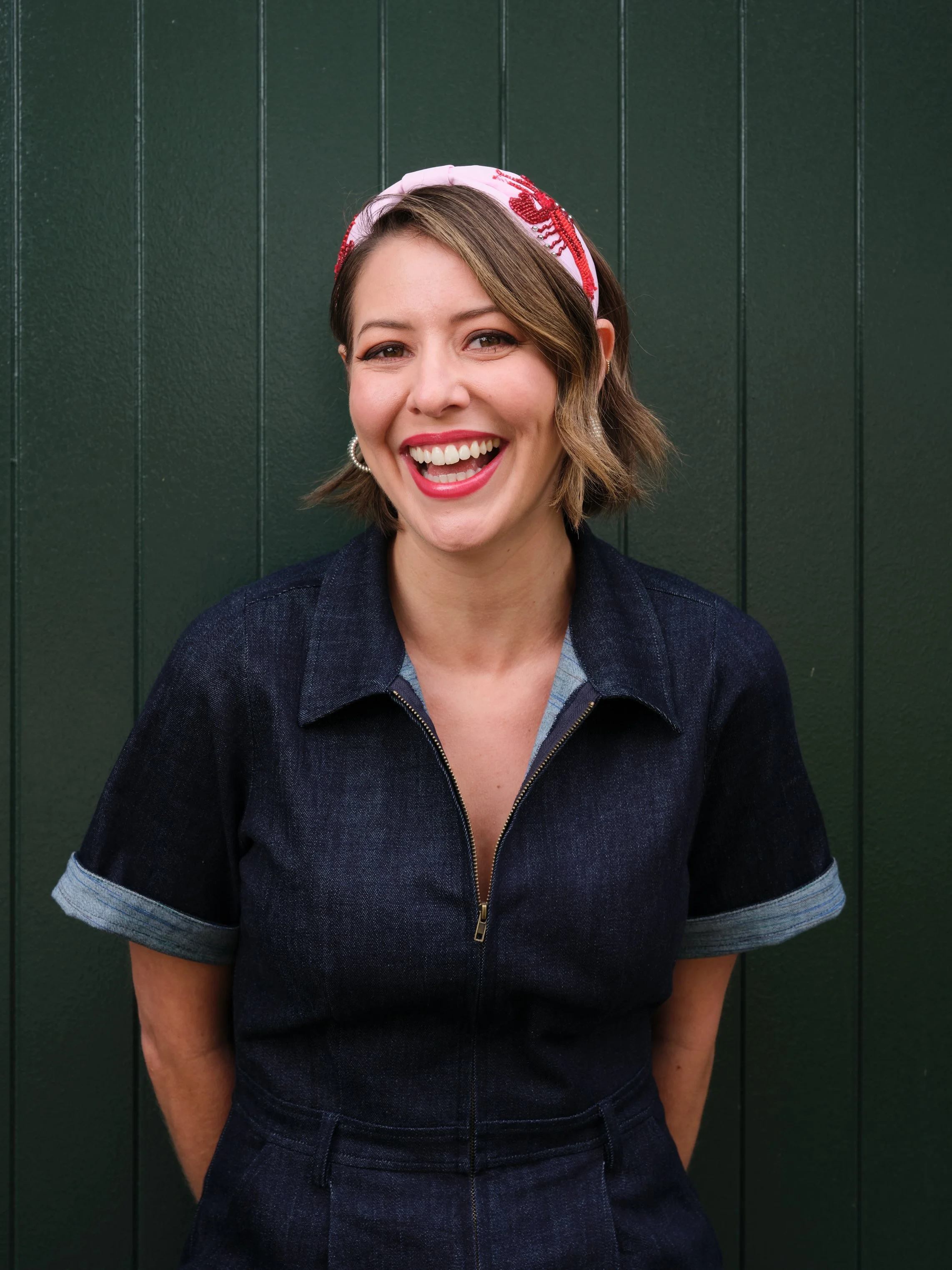 A woman with short brown hair, wearing a pink and red headscarf, a dark denim jumpsuit, and earrings, smiling in front of a dark green wooden wall.