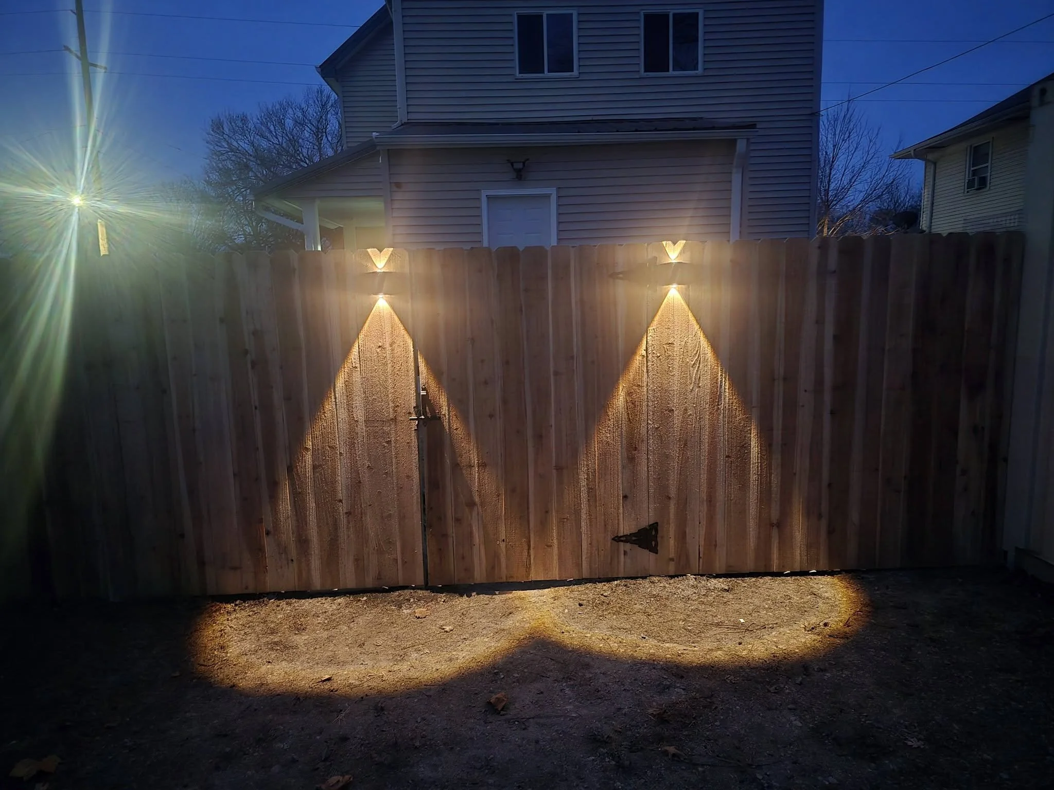 A wooden fence illuminated by outdoor lighting at night, with a two-story house in the background and bare trees against a dark blue sky.