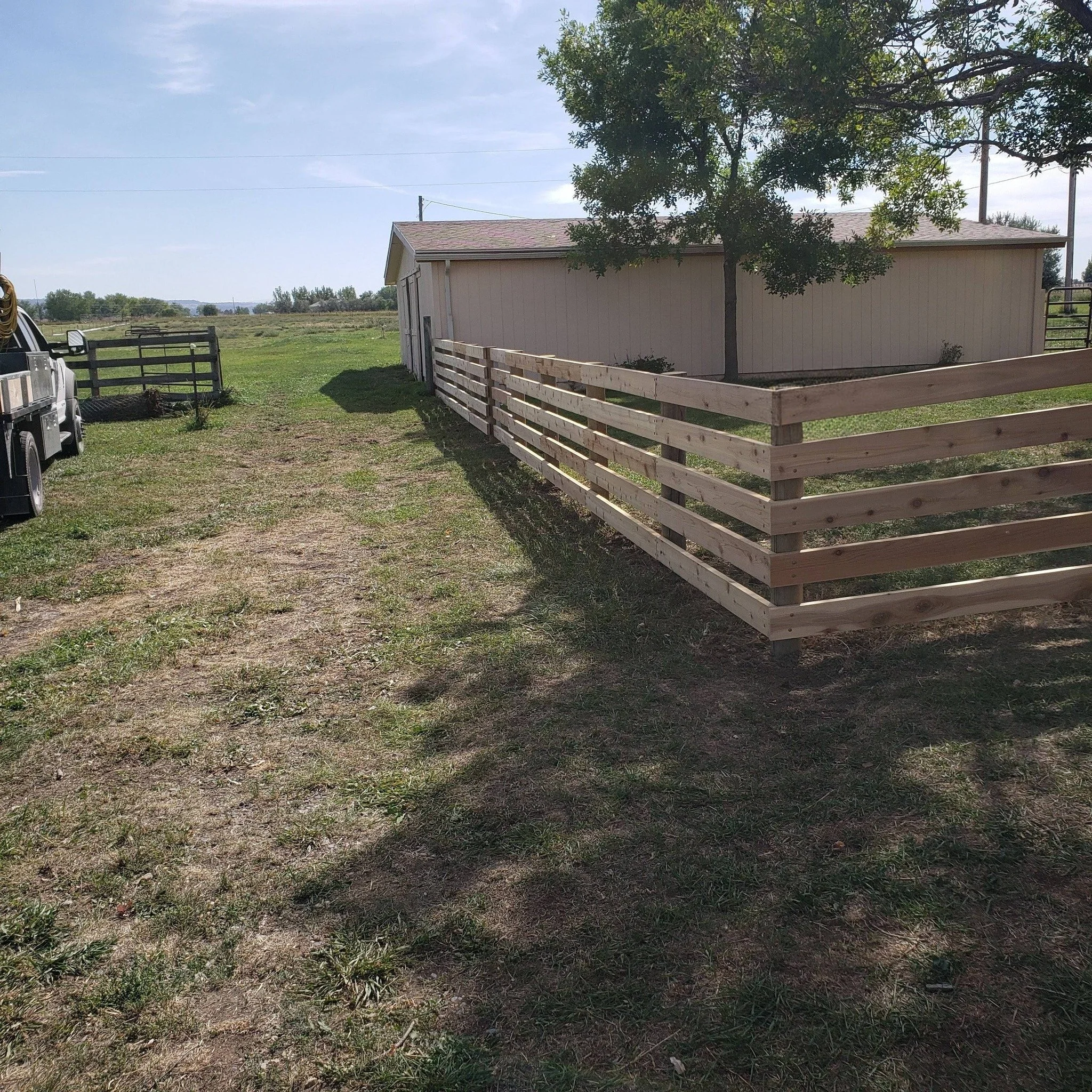 A rural scene with a white shed, a tree with green foliage, a wooden fence, a gravel pathway, and open fields under a blue sky with some clouds.