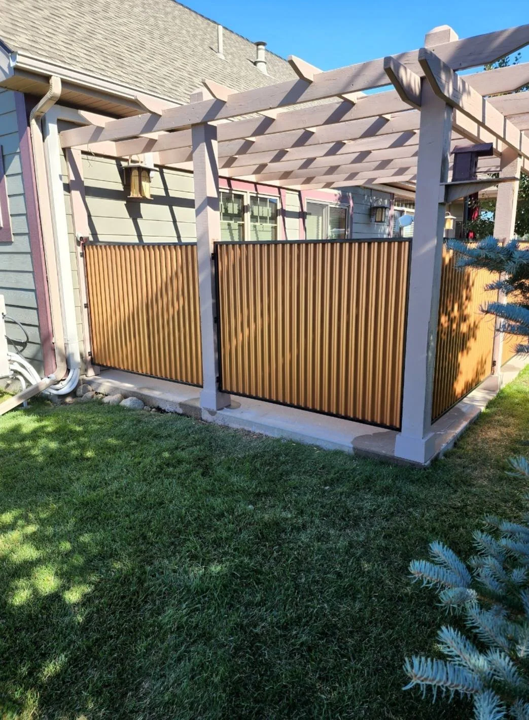 A backyard patio with a wooden pergola structure and matching brown privacy fence panels, adjacent to a blue house with windows and exterior lights, on a sunny day with green grass in the foreground.