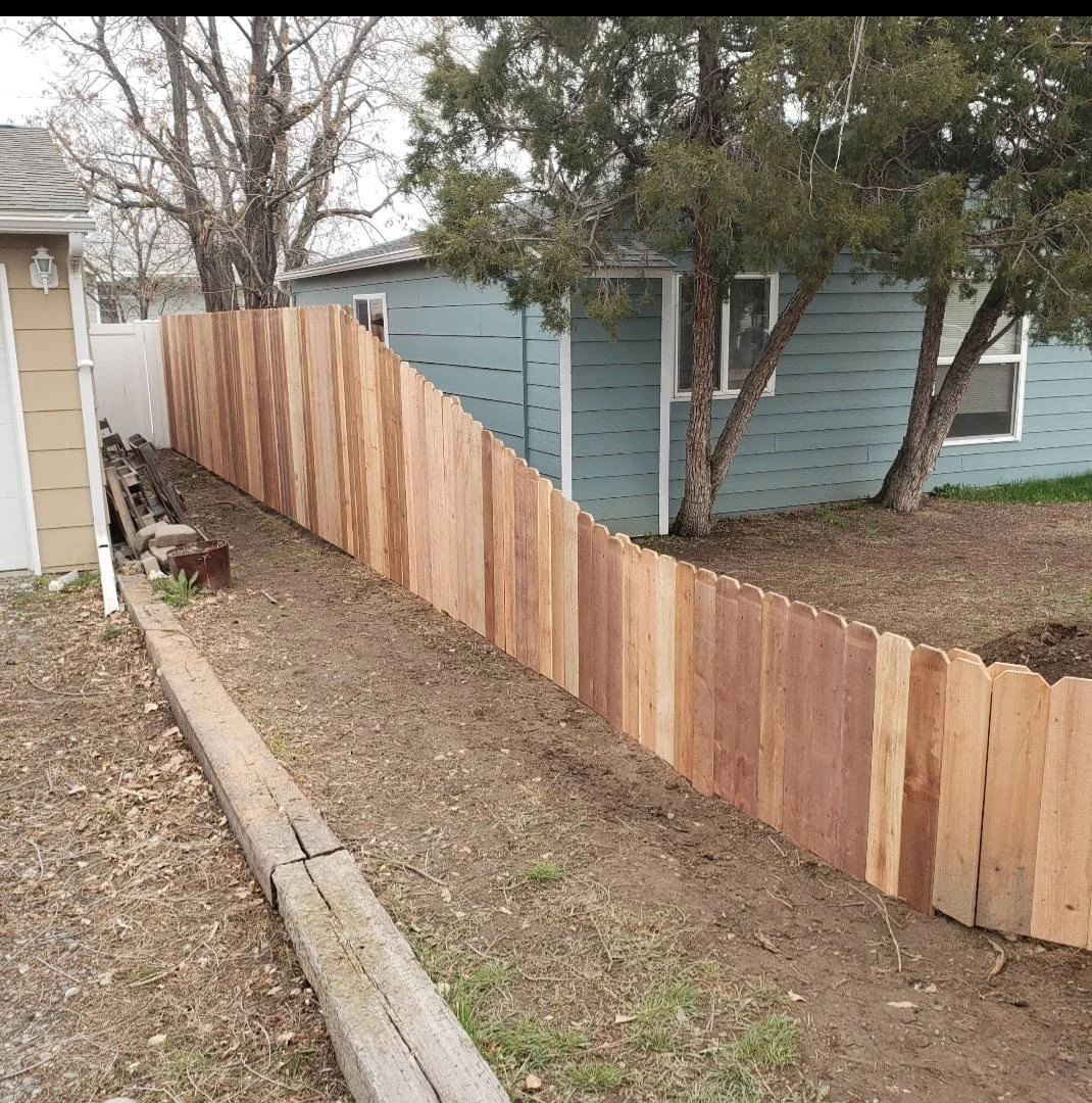 A newly built privacy wooden fence with scalloped edges in a backyard, adjacent to a blue house with trees nearby.