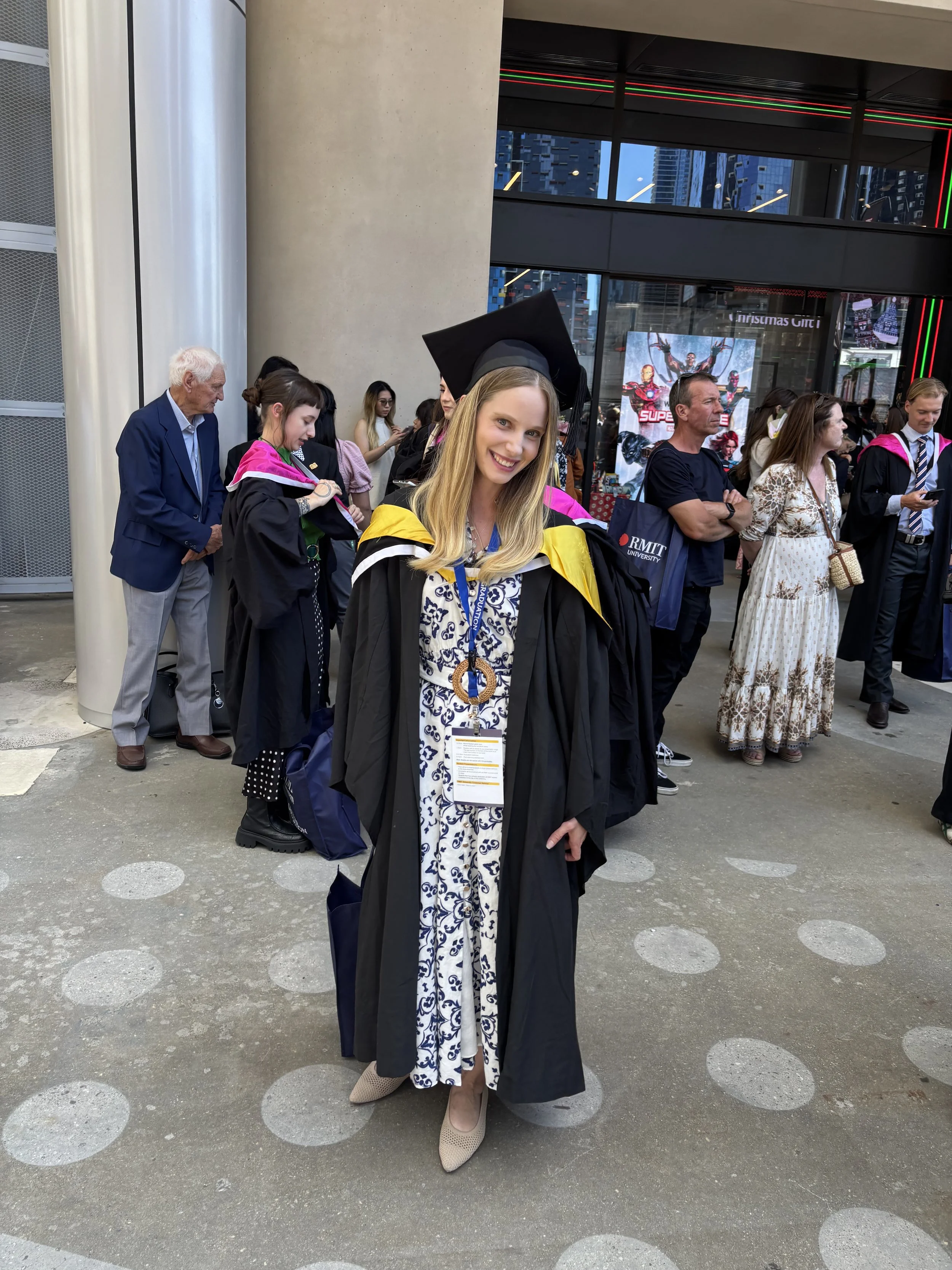 A young woman in graduation attire standing outdoors in front of a crowd, smiling at the camera, with other graduates and people in line behind her.