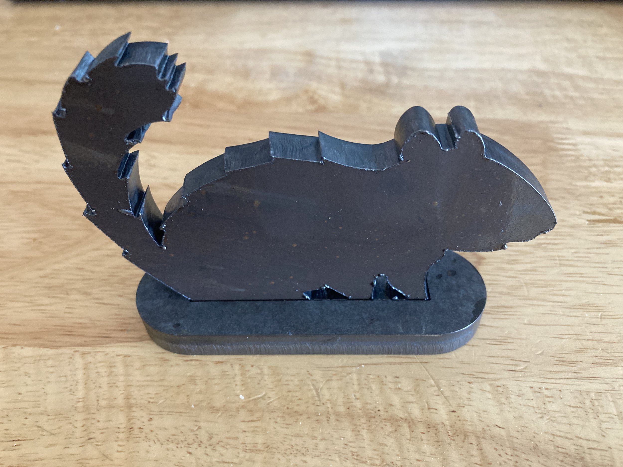 Black metal squirrel-shaped object with a bushy tail, standing on a flat base, placed on a wooden surface.