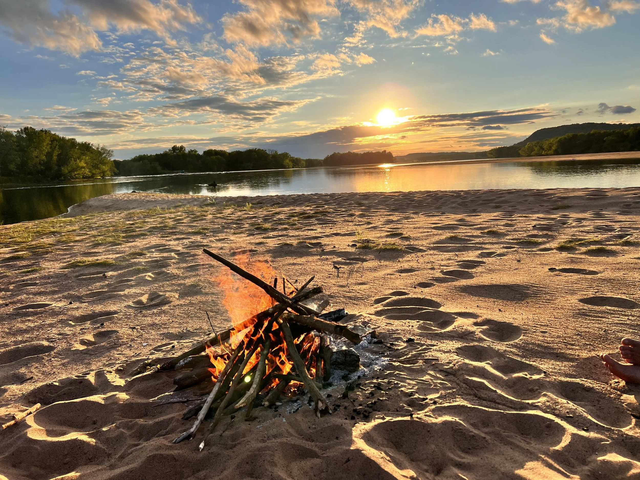 Sunset over a river on a sandy beach with a small campfire in the foreground and trees in the background.
