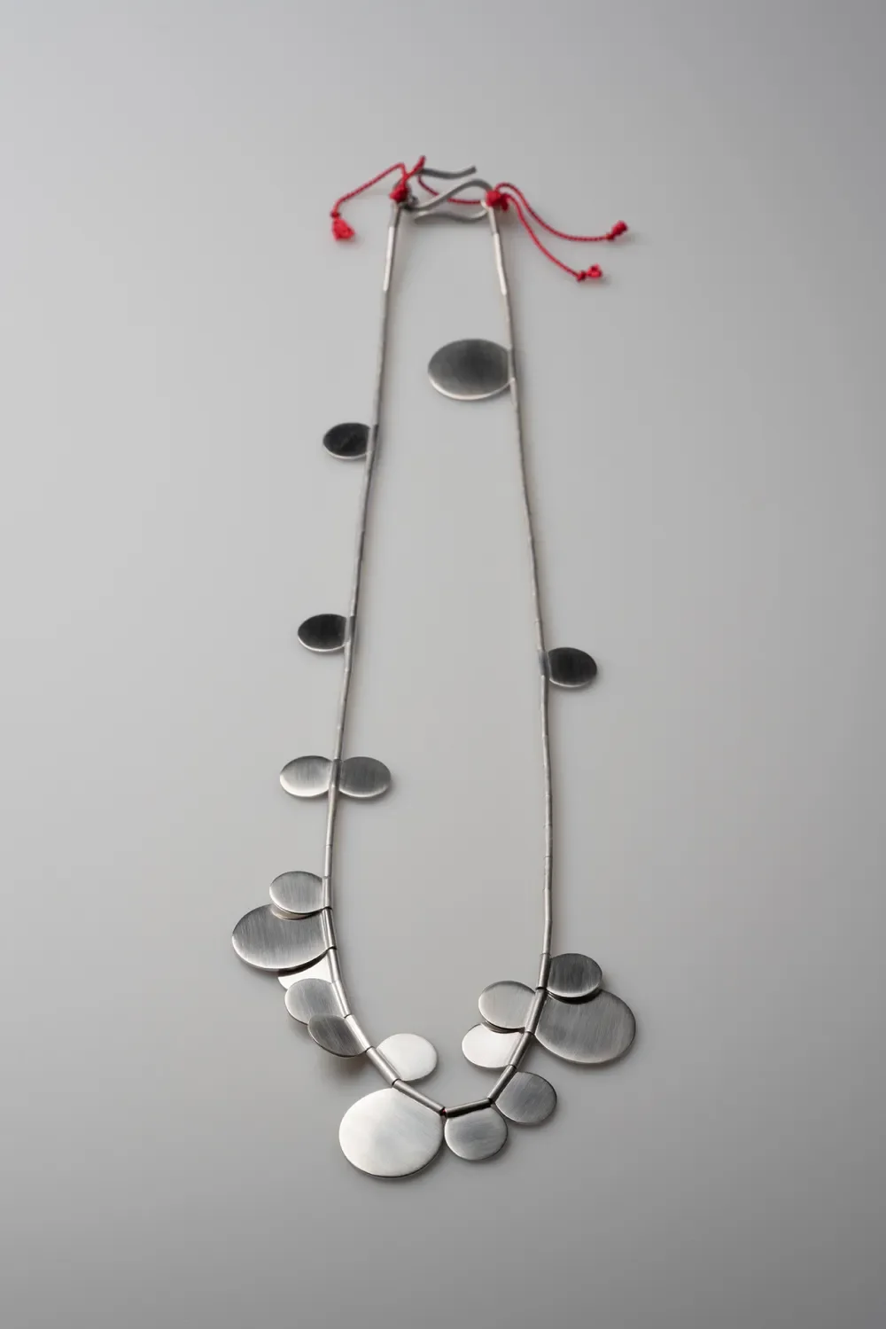A necklace with large, flat, metallic, oval-shaped pendants arranged in a graduated pattern on a metallic chain, attached with red string ties.