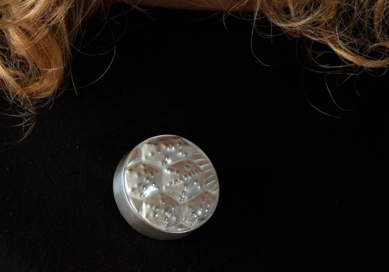 Close-up of a silver, round, textured object with small beads, placed on a black surface, with strands of blonde hair in the background.
