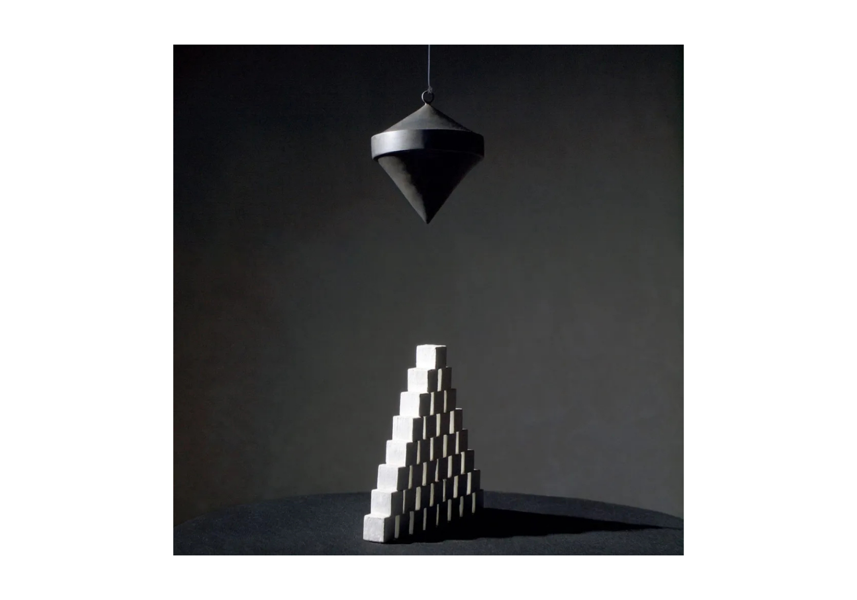 A construction of a pyramid made of small rectangular white blocks with a black rubber cone hanging above it, in a dark room with a black background.