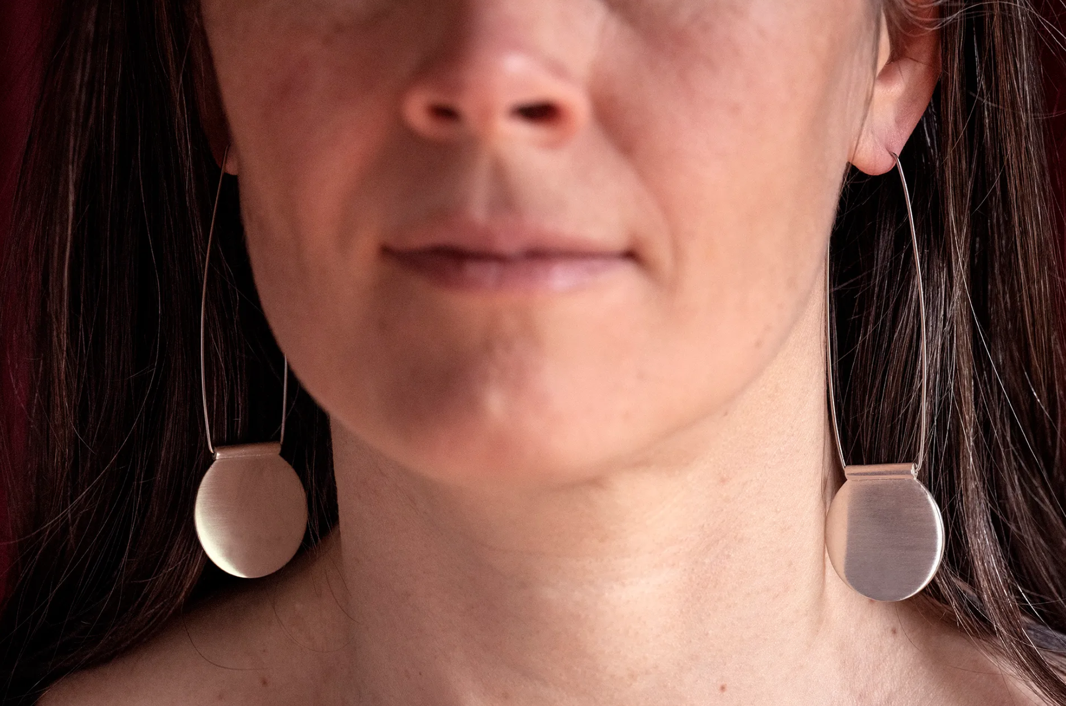 A close-up of a woman's face showing her lips, chin, and part of her nose and cheeks. She has long dark hair and is wearing large, round, metallic earrings.
