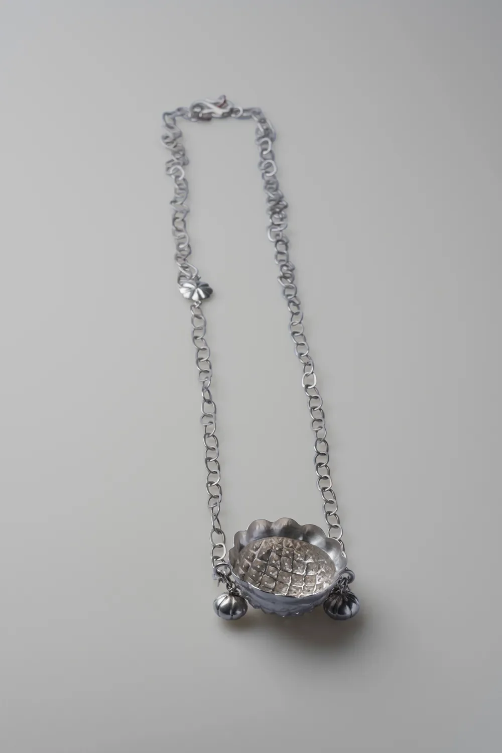 Silver necklace with a scalloped bowl pendant and two small beads on the chain, lying on a light gray surface.