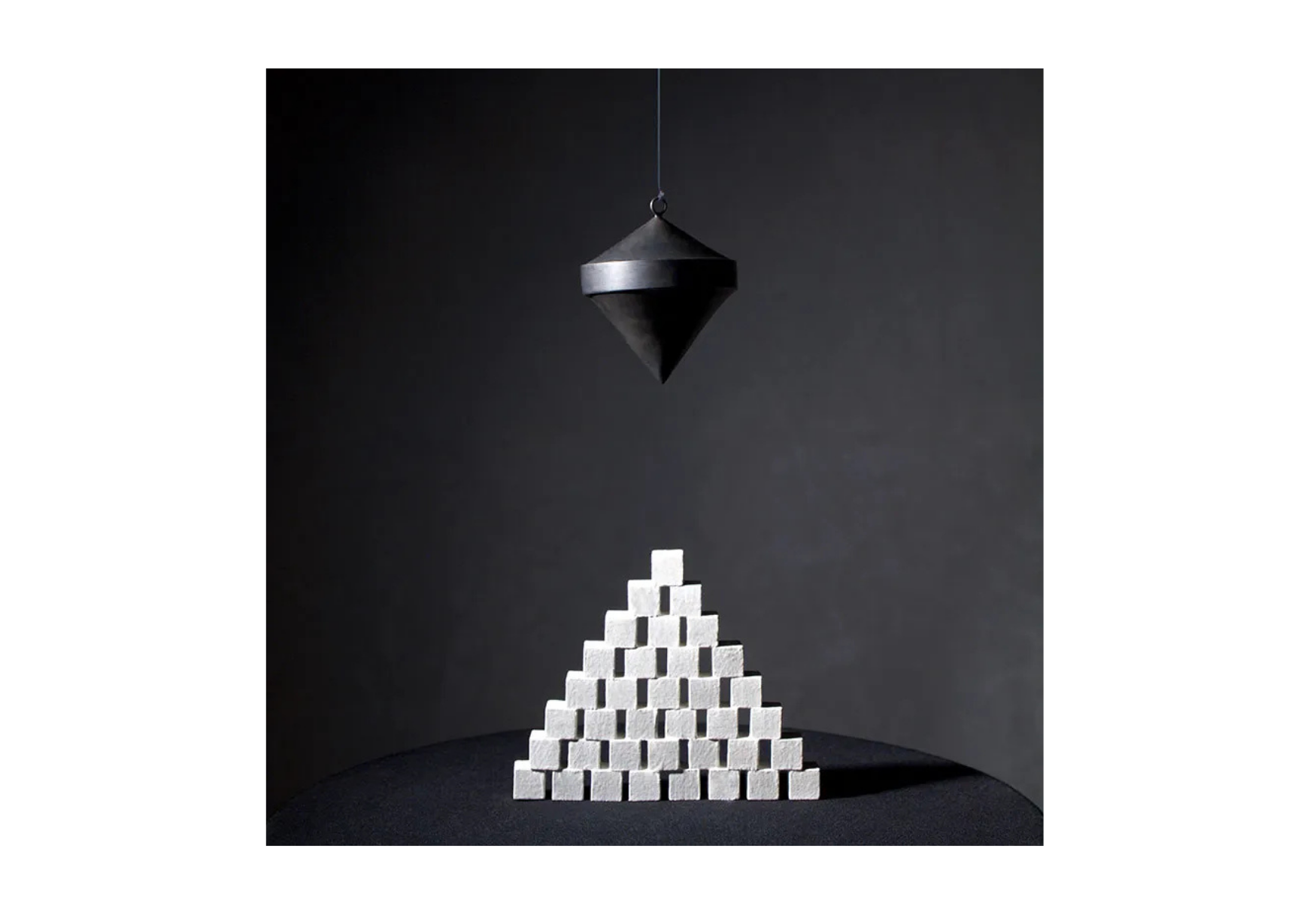 A black pyramid-shaped object hanging above a pyramid of white bricks against a dark background.
