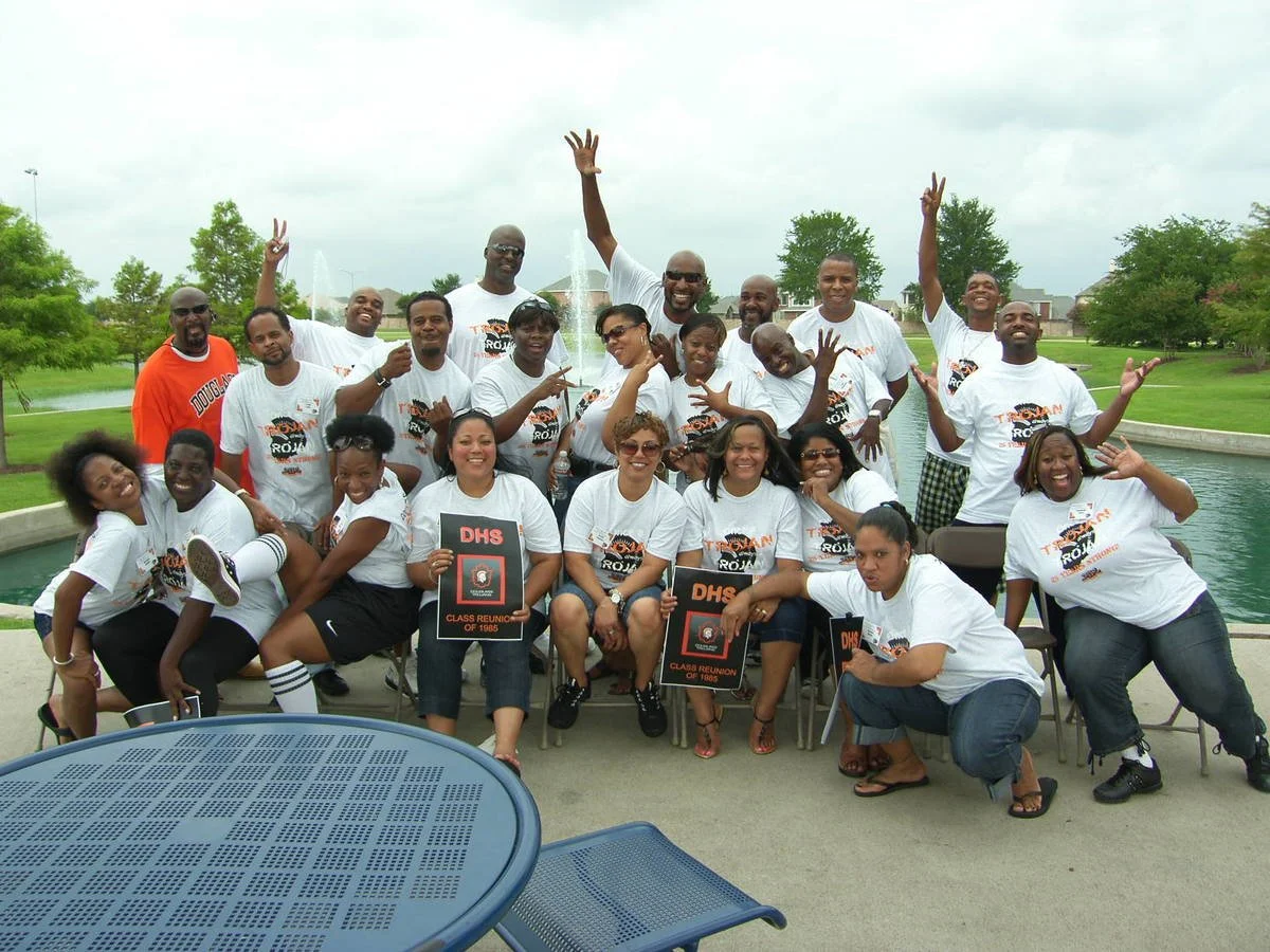 Group of people at a class reunion, some holding signs that read 'DHS Class Reunion of 1985,' posing outdoors near a fountain with trees and grass in the background.