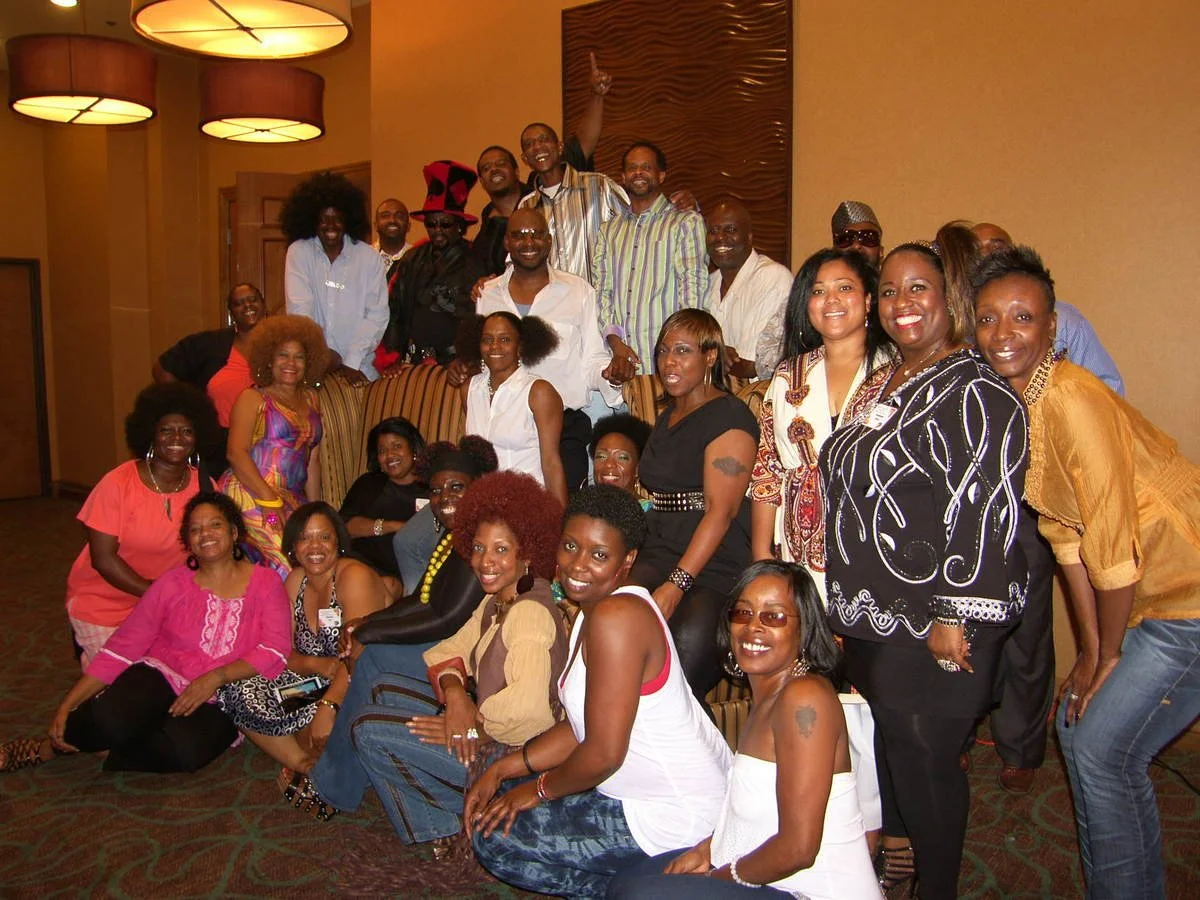 A large group of smiling people posing together in a room, some standing and some sitting, with warm lighting and a decorative wooden wall in the background.