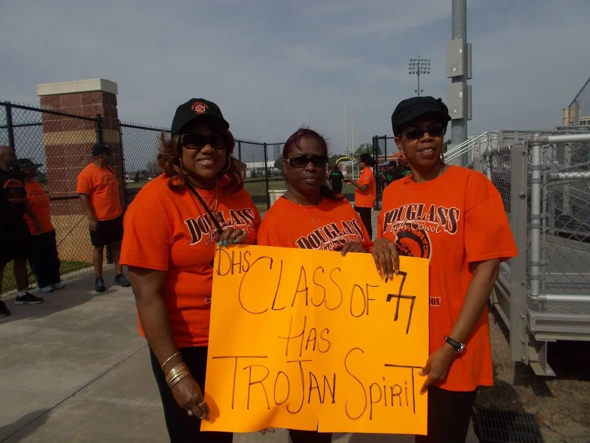 Three women wearing orange shirts with 'Douglas High School' printed on them, holding a yellow sign that reads 'DHS Class of 77 has Trojan Spirit' at a school event outside football field.