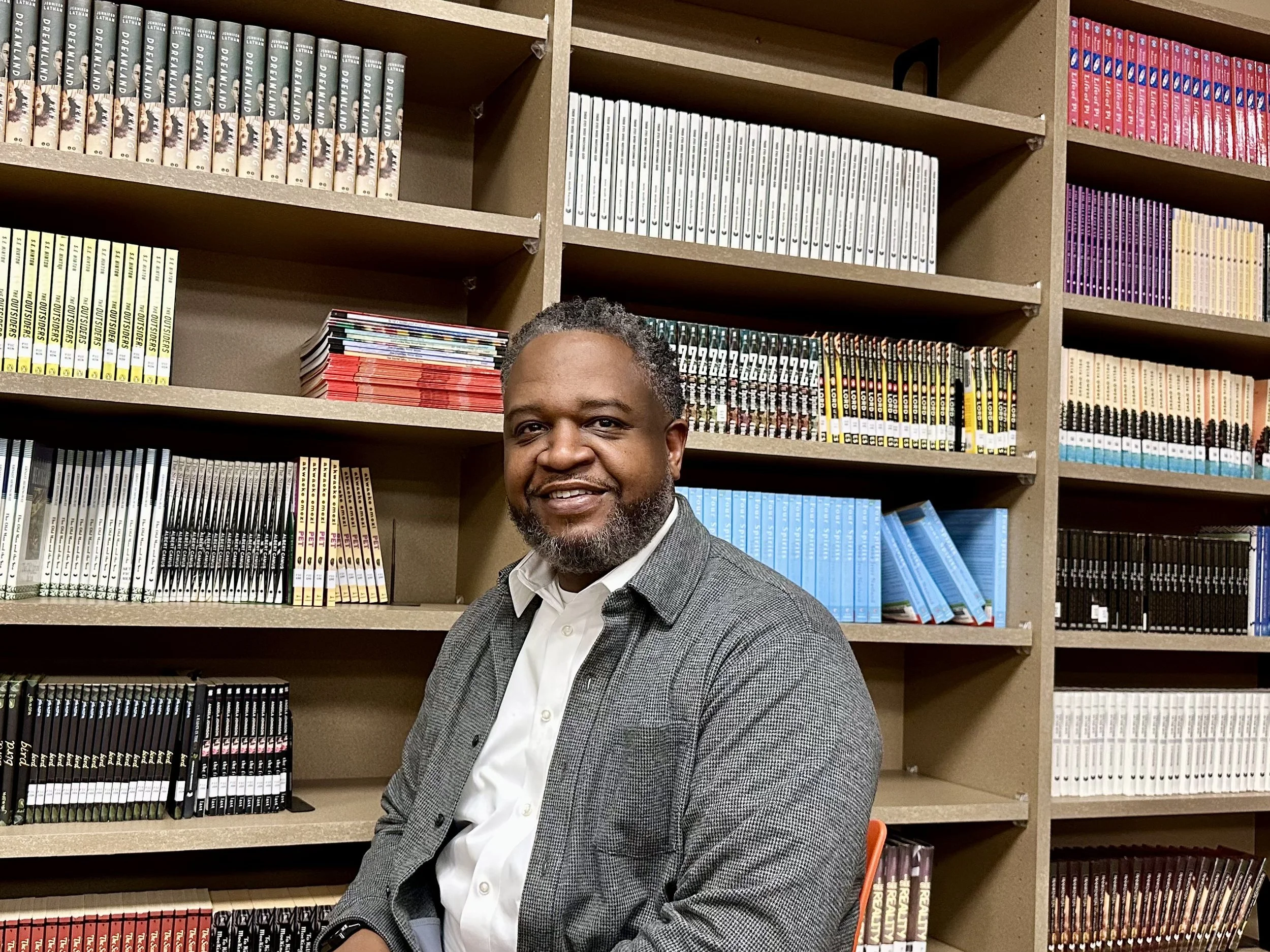 A man sitting in front of bookshelves filled with manga, graphic novels, or comic books in a library or bookstore.
