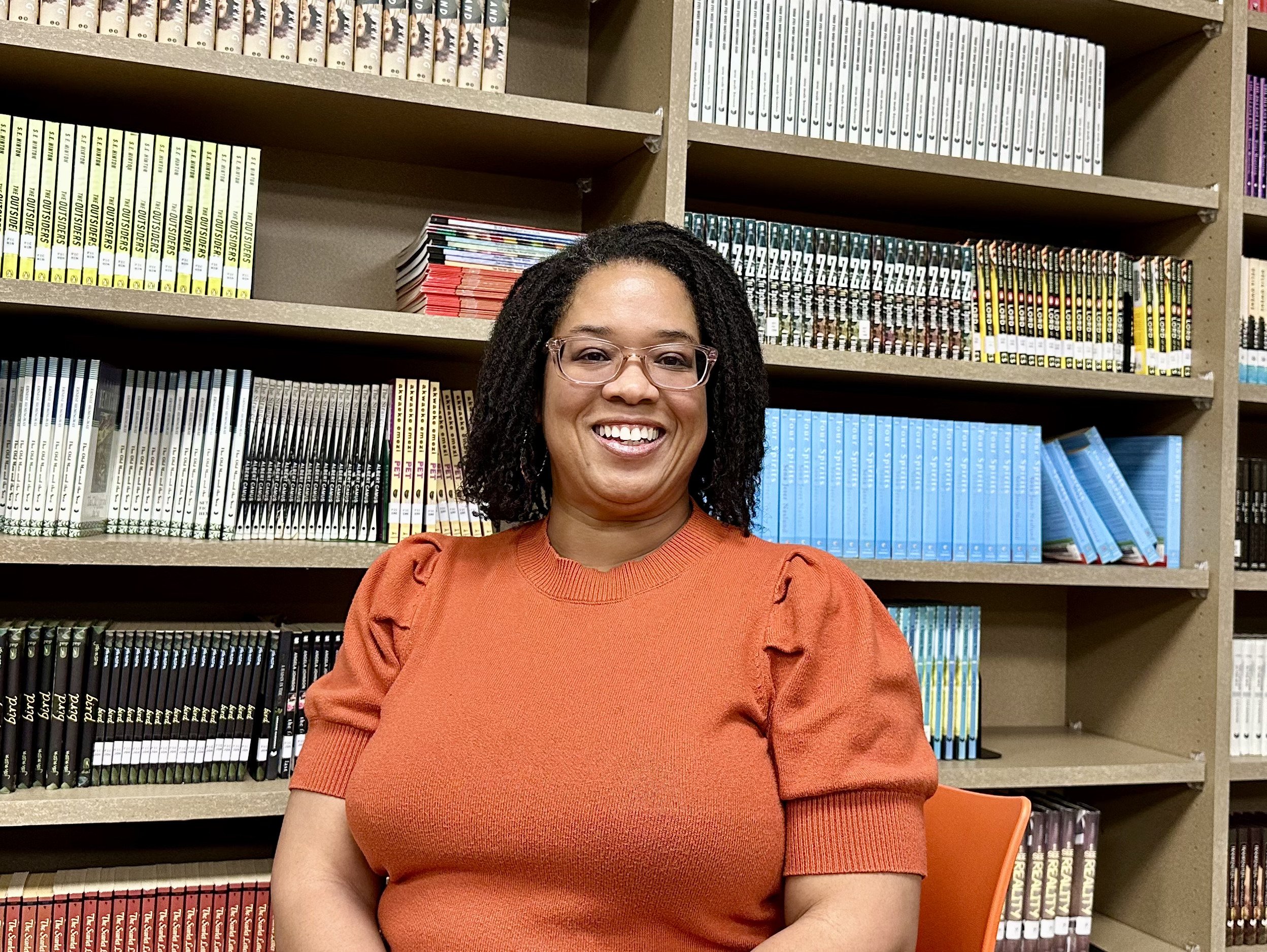 A woman wearing glasses and an orange top sitting in front of shelves filled with books.