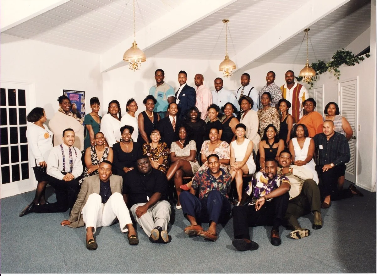 Group of diverse adults gathered in an indoor venue, posing for a photo with some smiling and sitting while others stand behind them.