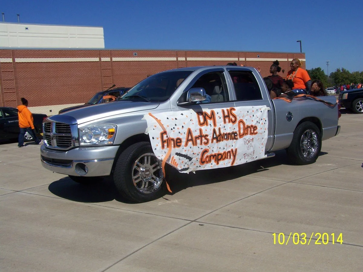 A silver pickup truck decorated for a parade with a large banner on the side that reads 'DM HS Fine Arts Advance Done Company,' carrying people in the truck bed. Other parade vehicles and people are visible in the background on a sunny day.