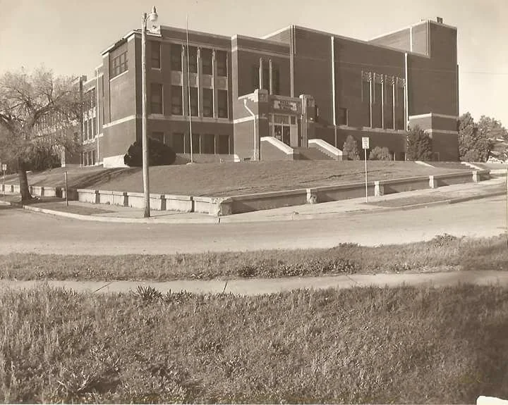 A black and white photo of a large brick school building on a hill with a staircase leading up to the entrance, surrounded by trees and a grassy area with a sidewalk in the foreground.