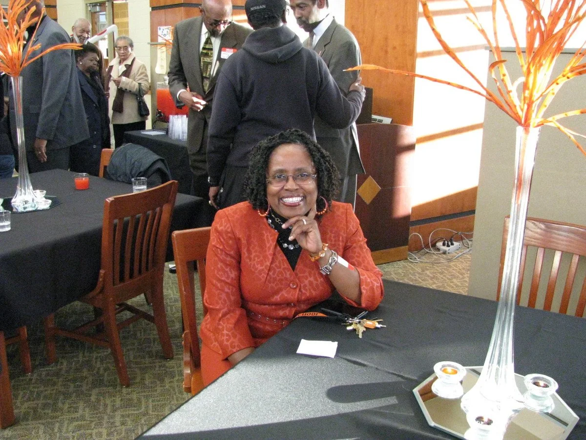 A woman wearing glasses and an orange outfit smiling and sitting at a dining table with a black tablecloth during a social event. There are flower arrangements, candles, and other people in the background, some engaged in conversation.