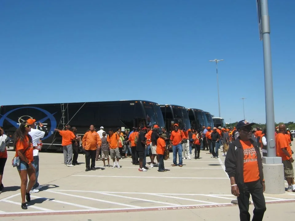 People wearing orange shirts and hats gather in a parking lot with buses parked behind them, under a clear blue sky.