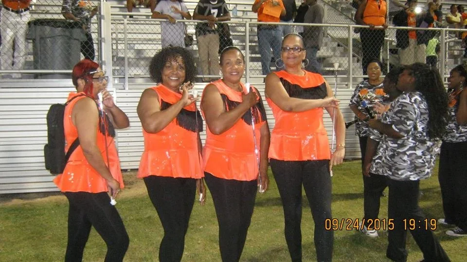 Four women standing on a field in front of bleachers, wearing coordinated orange and black outfits, smiling and waving at the camera during an outdoor event at night.