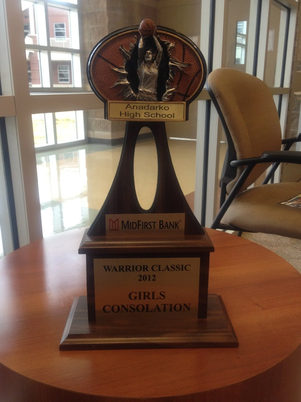A basketball trophy with a female figure dunking a basketball, surrounded by flames, on a wooden base. The base has plaques that read 'Anadarko High School,' 'MidFirst Bank,' and 'Warrior Classic 2012 Girls Consolation.'