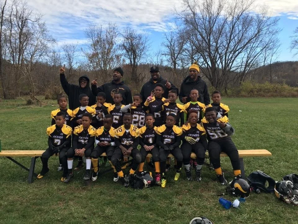 A youth football team called the Vikings, dressed in black and yellow uniforms, posing with their coaches on a grassy field with trees in the background. The team members are sitting and standing, and some are making celebratory gestures.