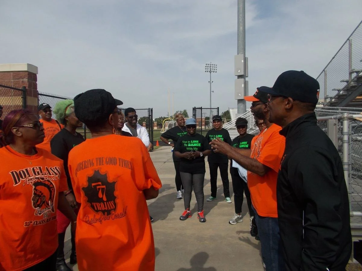 Group of people gathered outdoors near a sports field, some wearing orange and black shirts, engaged in conversation or listening to a man speaking.