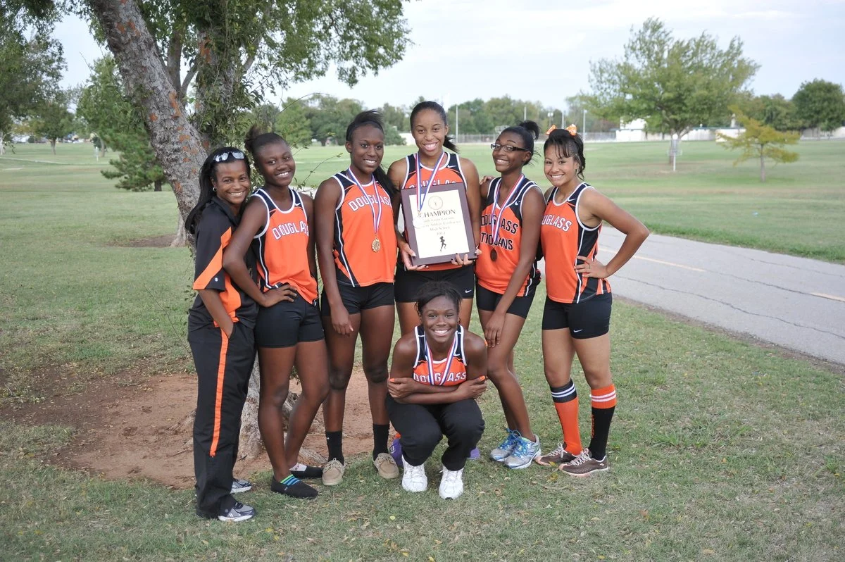 Group of six young female athletes in orange and black sports uniforms posing for a photo on a grassy field, one holding a framed championship plaque, and another with a medal around her neck.