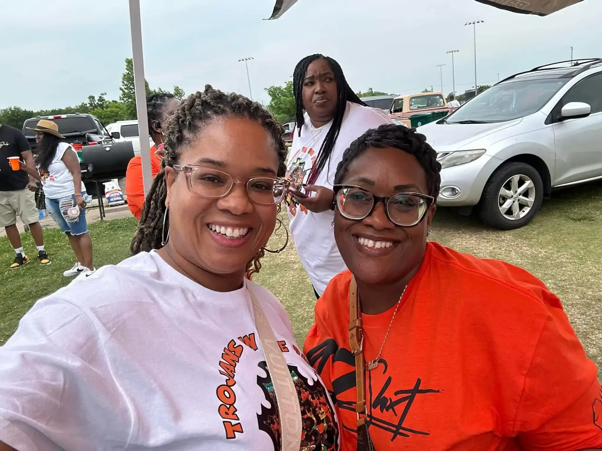 Three women at an outdoor event smiling at the camera with cars and other people in the background.