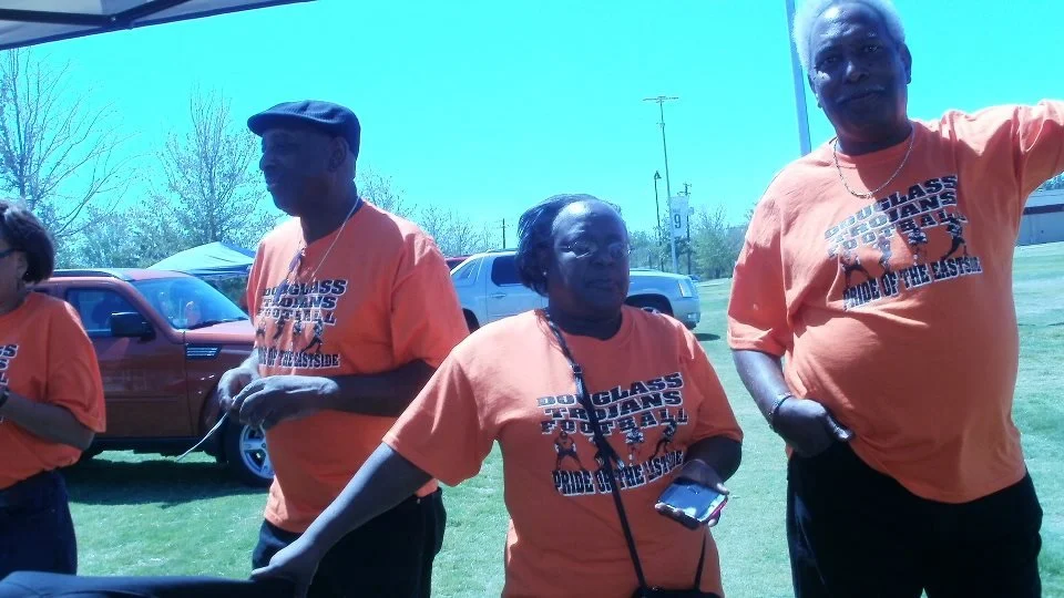 Three people wearing matching orange T-shirts attending an outdoor event, parked cars and trees in the background.