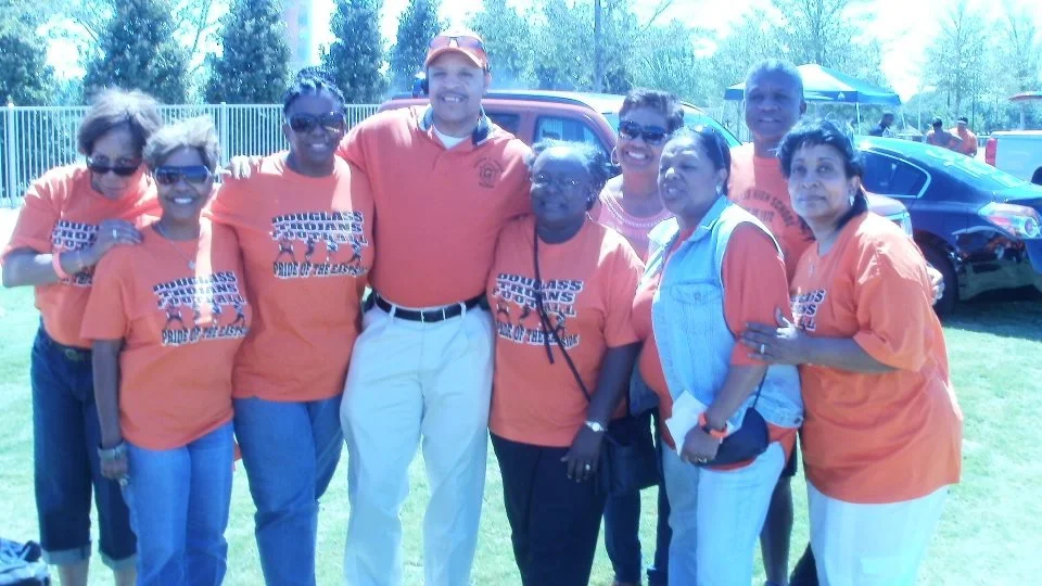 Group of nine people standing outdoors on grass in front of cars, all wearing orange T-shirts with a logo and text, smiling at the camera.