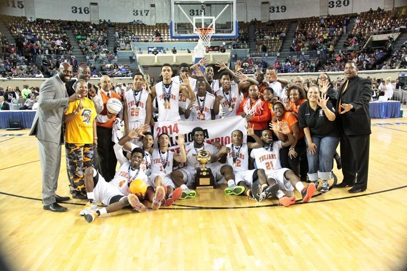A youth basketball team celebrating on the court with trophies and medals after winning a championship, surrounded by coaches and supporters.