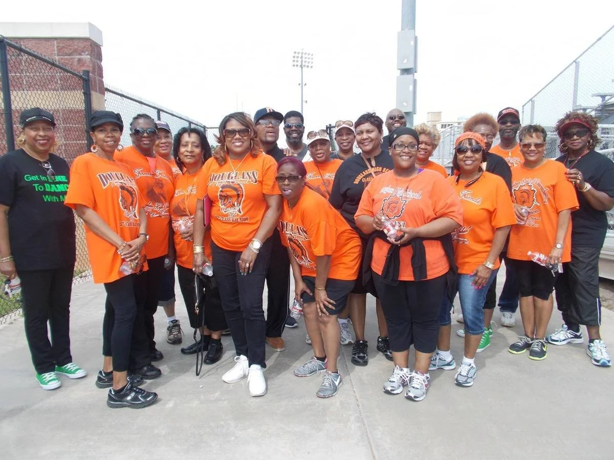 Group of women wearing orange T-shirts with a lion logo, standing outdoors near a chain-link fence, smiling and posing for the photo.