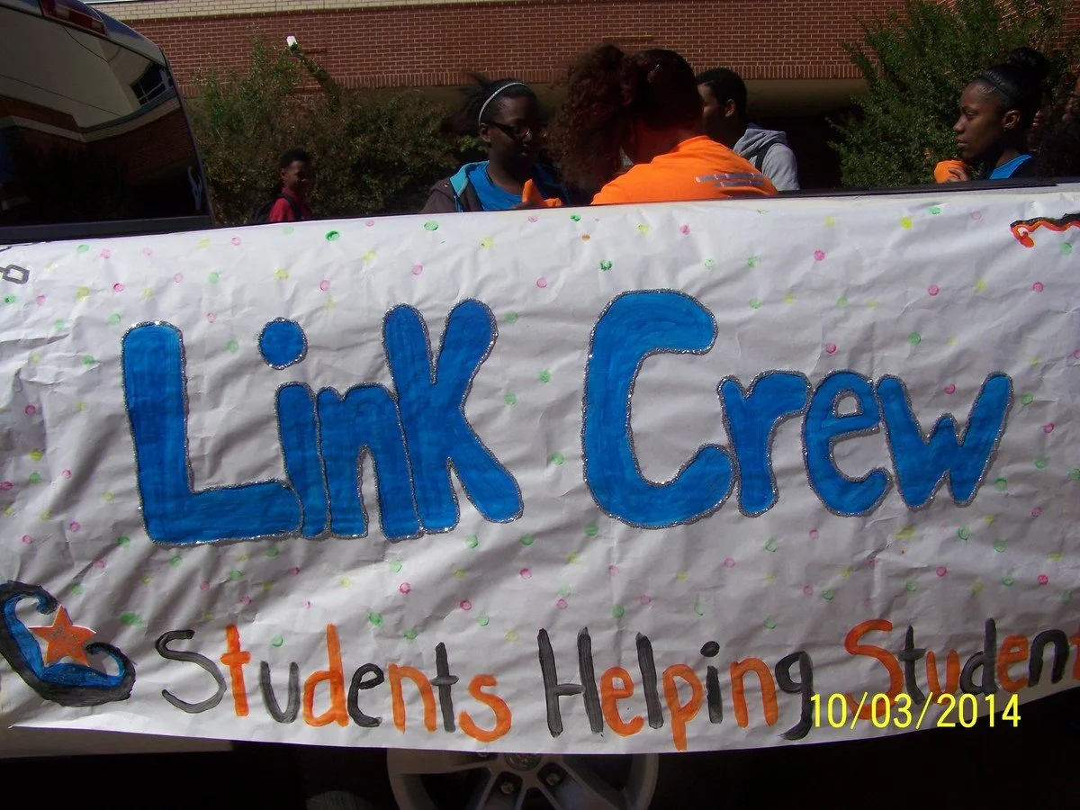 A group of students holding a banner that says "Link Crew" and "Students Helping Students" at an outdoor event, with a brick building and bushes in the background, dated 10/03/2014.