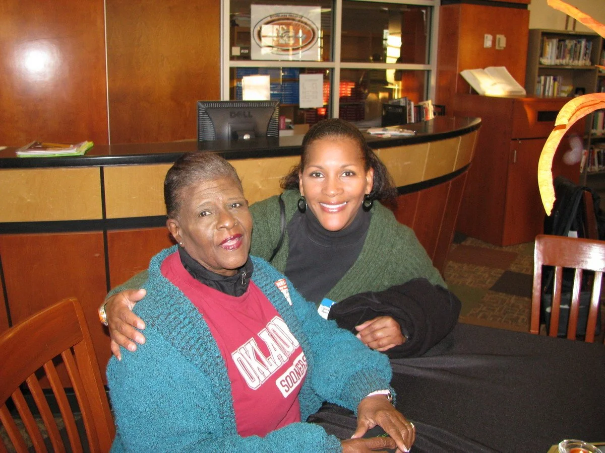 Two women are sitting at a table in a library, smiling and posing for the photo. One woman is older, wearing a red Oklahoma Sooners shirt and a blue cardigan. The other woman is younger, wearing a black top and green sweater. There are bookshelves an