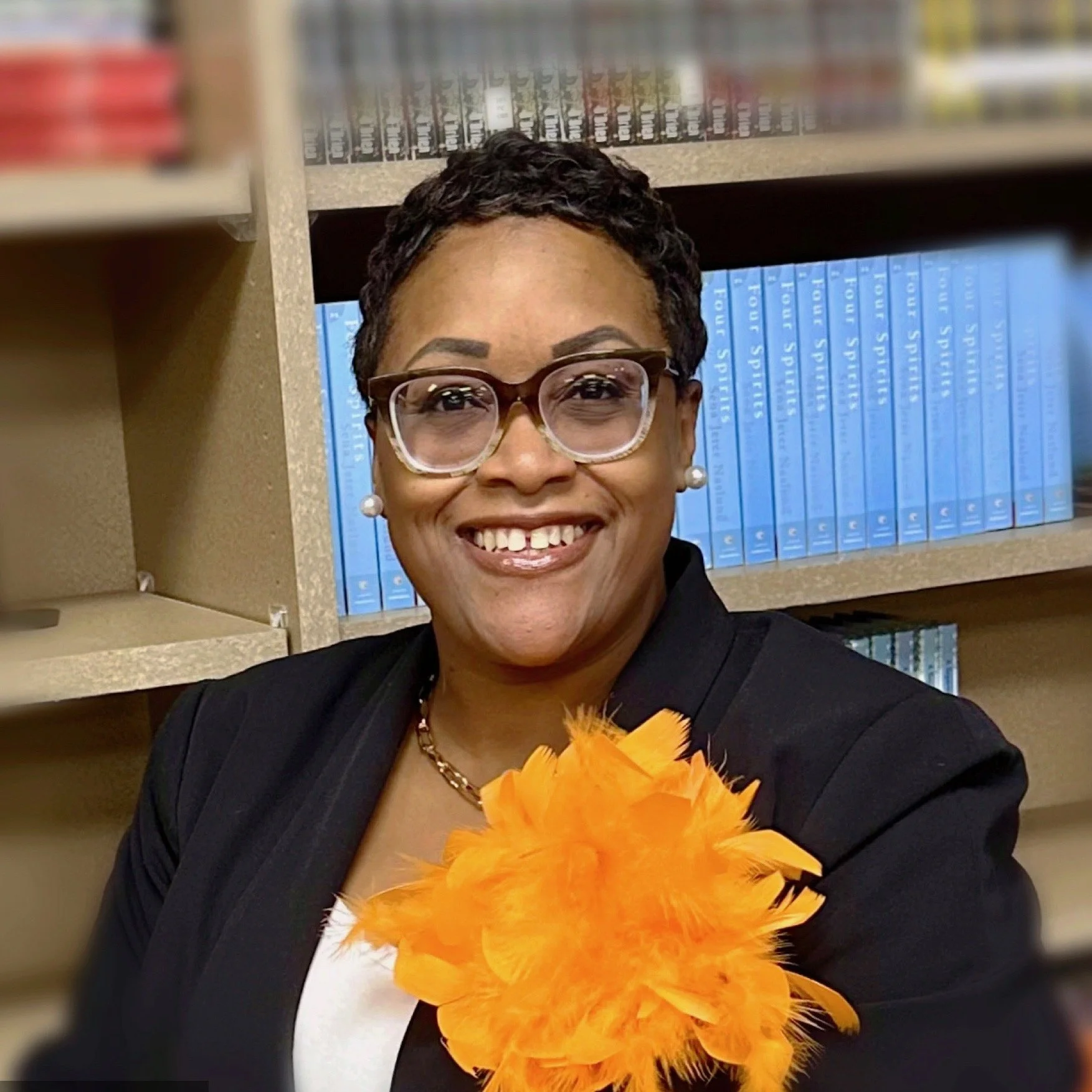 A smiling woman with short curly hair, glasses, pearl earrings, and a black blazer, holding an orange feathered accessory, standing in front of a bookshelf filled with blue books.