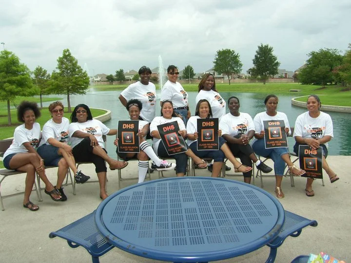 A group of women holding certificates and wearing matching white shirts, sitting and standing by a pond in a park.