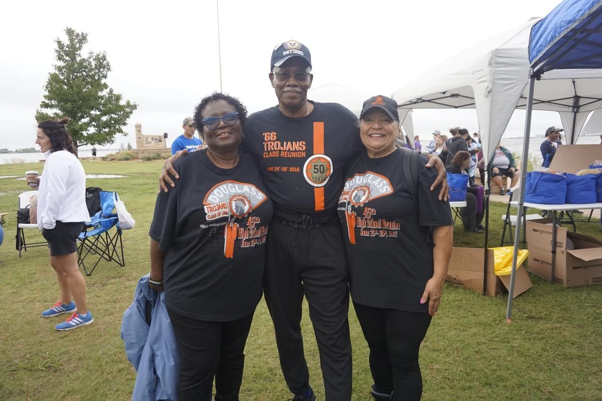 Three people, two women and one man, stand together outdoors at a park during a community event. They are all wearing black T-shirts with orange and white graphics celebrating their high school reunion and anniversary. The man in the middle is wearin