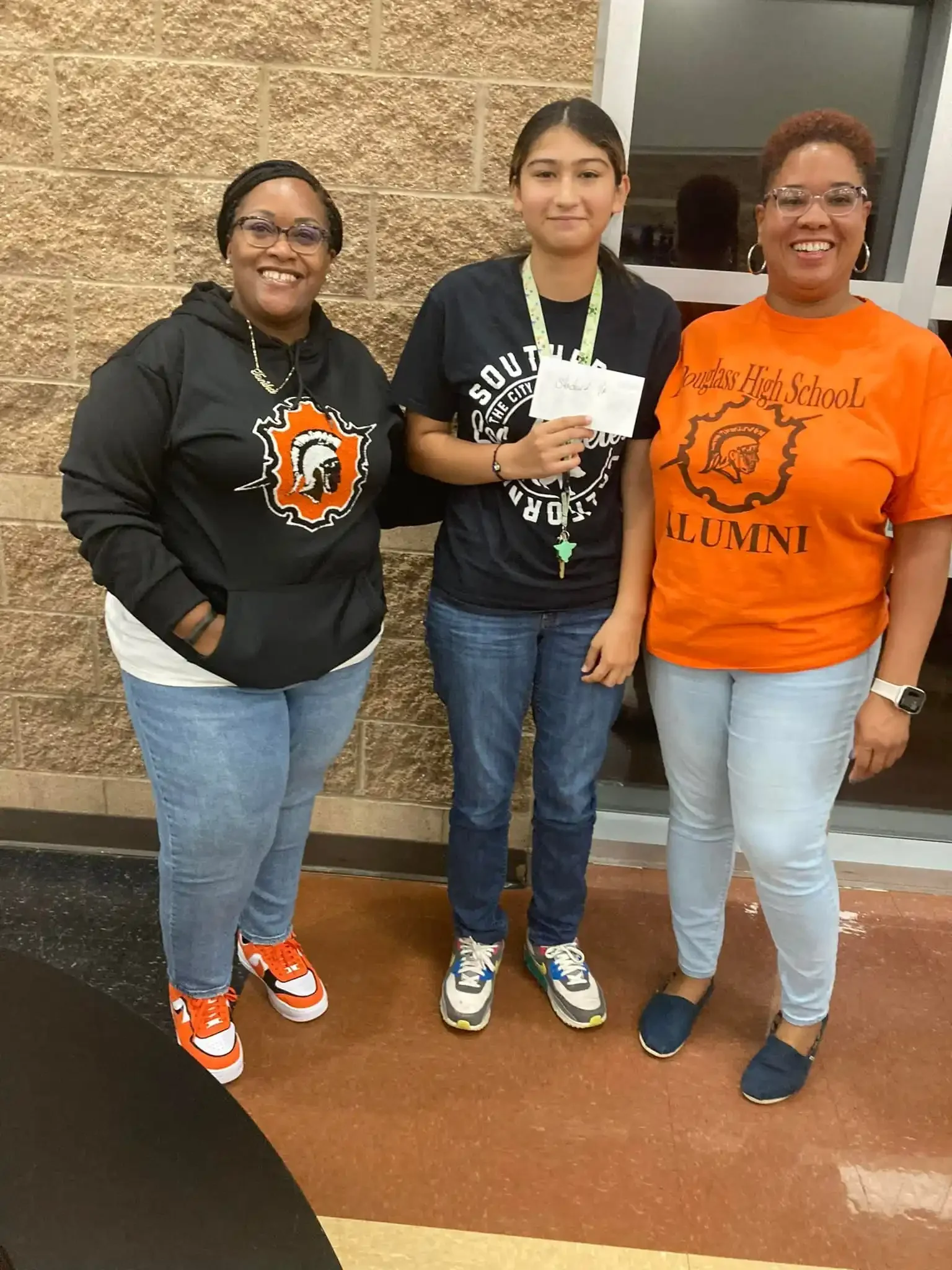 Three women and a young girl standing together indoors, smiling, with the girl holding a small paper and wearing an 'South High School' shirt.