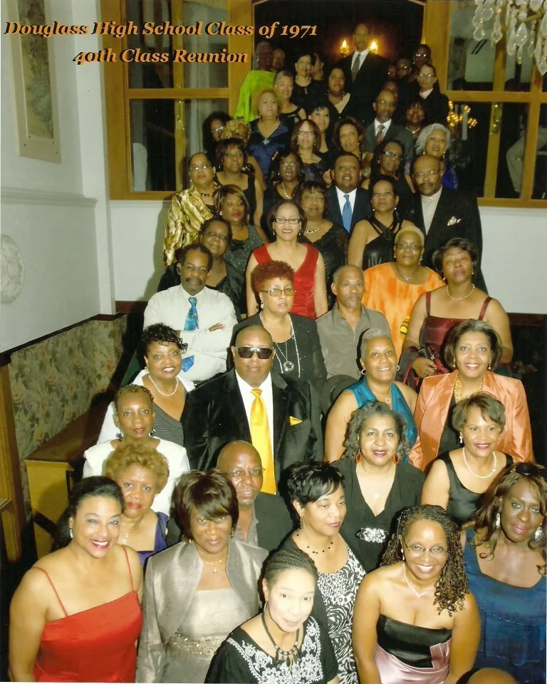 Group of mostly African American adults gathered indoors for a class reunion. The event commemorates the Douglass High School Class of 1971's 40th reunion. The group is dressed formally, with women in dresses and men in suits or dress shirts. The bac