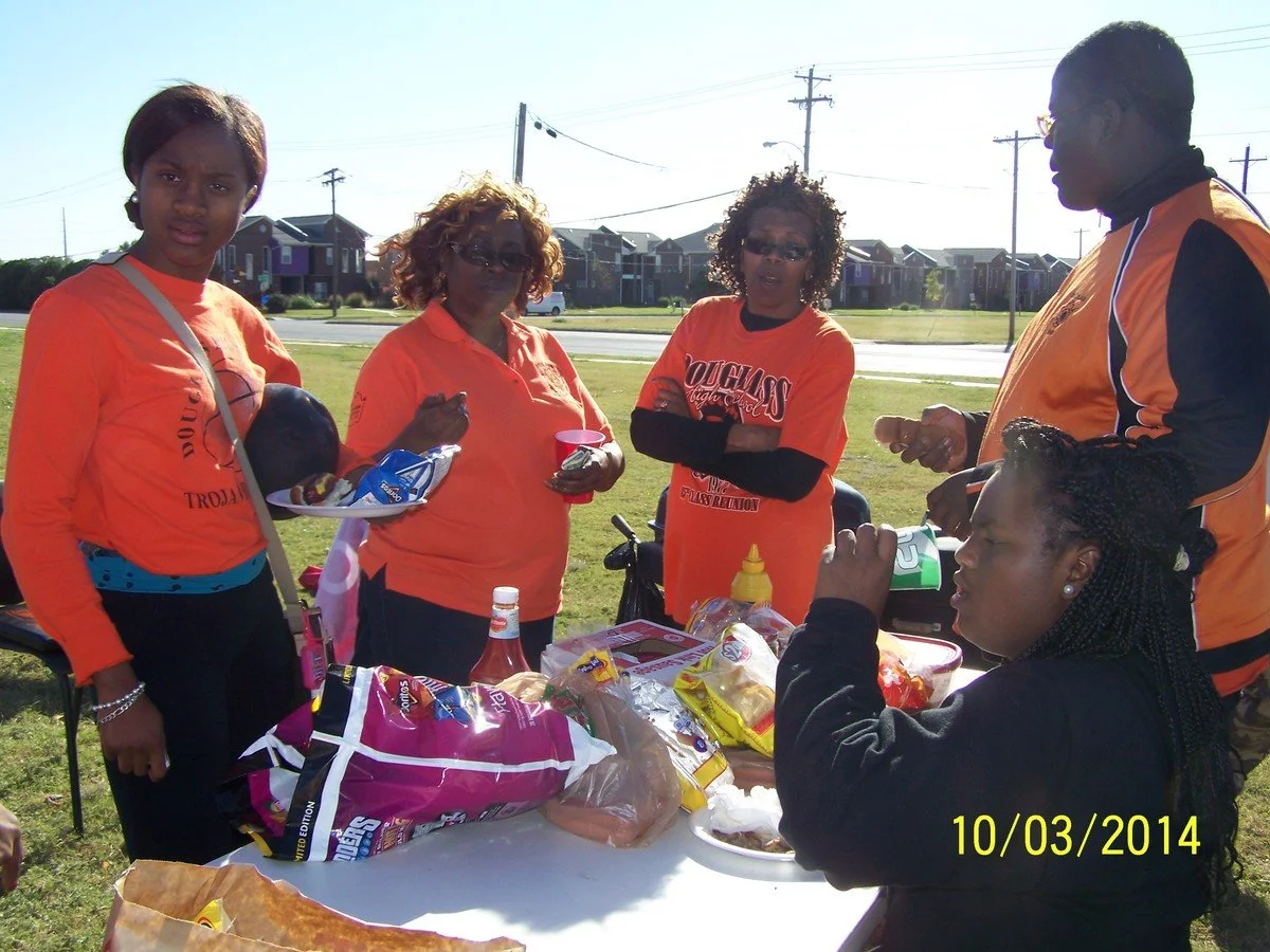 Six women gathered outdoors at a table with food, wearing matching orange shirts, in a grassy area near residential houses and power lines, on a sunny day.